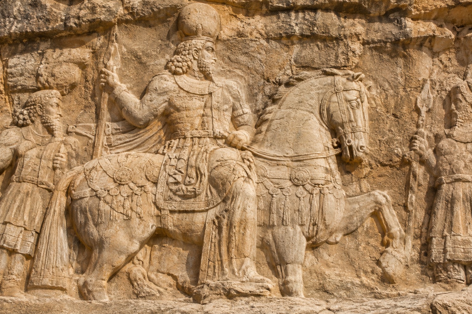 Illustration d’un relief rocheux d’inspiration sassanide, représentant un souverain couronné à cheval entouré de personnages dans une pierre ocre.